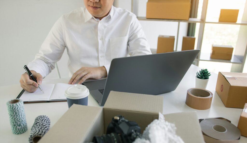 Young asian man looking on computer laptop and taking writing order list on paper