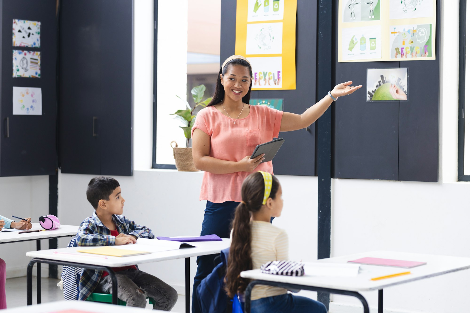 A young biracial female teacher points at posters, holding tablet, in a pink top